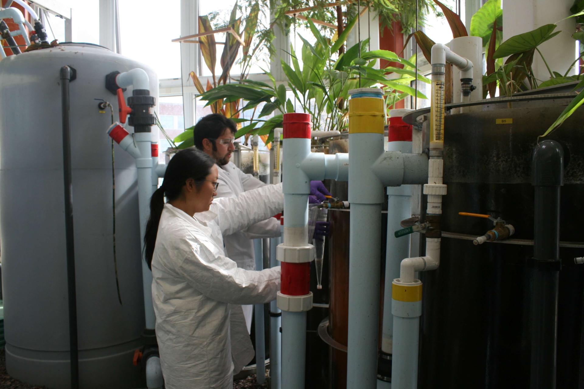 Two students performing Lab work next to large tanks of water with tubing attached.