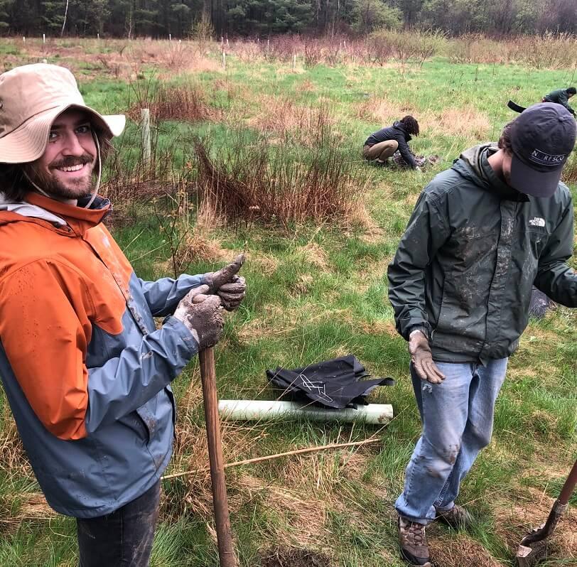 Students on a rainy day digging holes for ecological restoration, one is smiling and giving a thumbs up to the camera