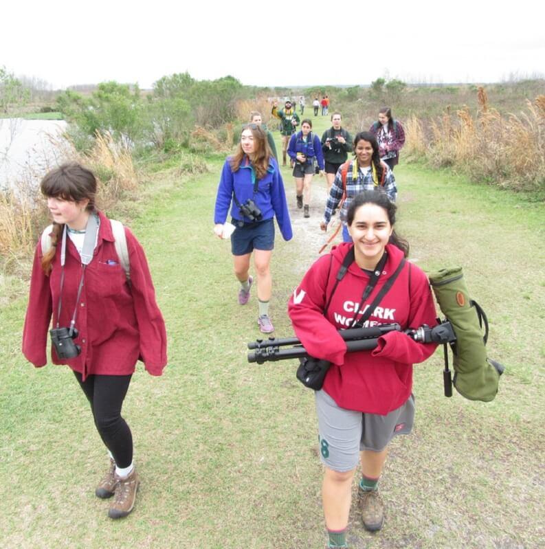 Students walking along a grassy shoreline holding binoculars and photography equipment
