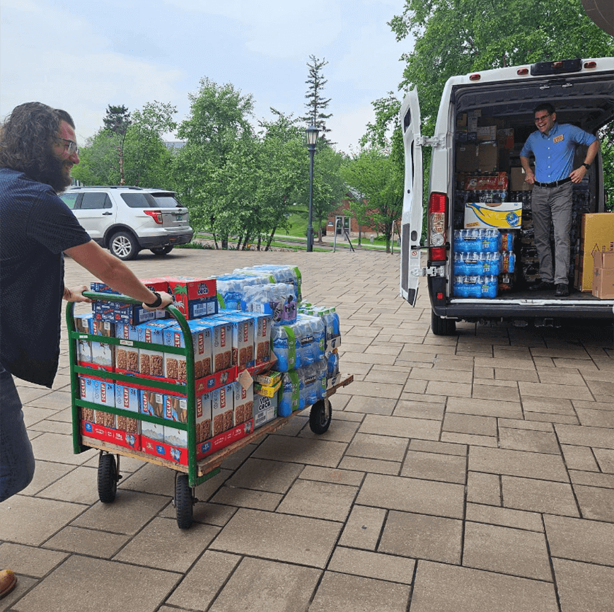 A person pushes a large cart full of cases of water bottles and granola bars toward an open van.